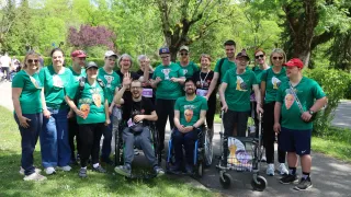 A group of people wearing green t-shirts with carrot designs pose for a photo. Some are in wheelchairs. They are standing on a grass path in a park. Behind them are trees and bushes.