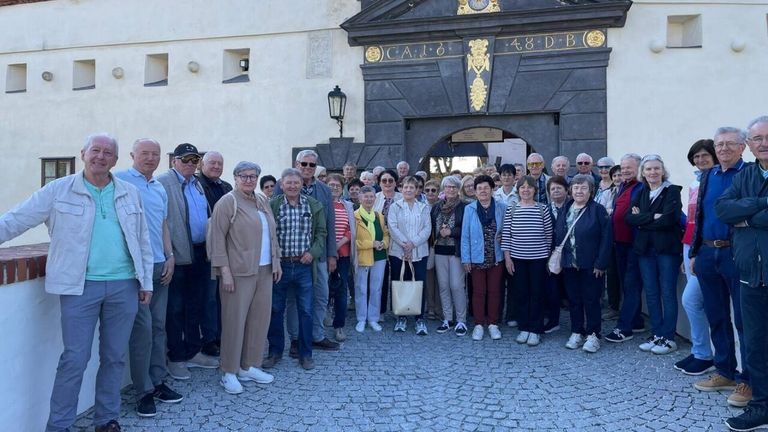 A group of elderly individuals wearing sunglasses and various outfits are standing in front of a stone building with an archway.