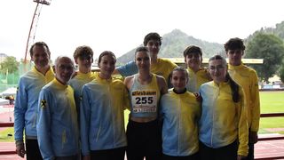 A group of athletes in yellow and blue uniforms, one woman in front wearing a number 255, standing on a track with mountains in the background.