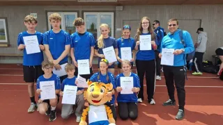 A group of young athletes, some holding certificates, are posing for a photo. A mascot in a tiger costume is in the center. Behind them, a building with glass windows and a roof is visible.