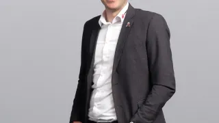 A man is standing with a smile on his face. He is wearing a white shirt and a black blazer. He has a watch on his left wrist and a logo on his blazer.