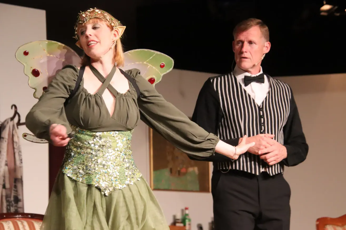 A woman in a green dress with fairy wings and a man in a striped suit hold hands on stage. The woman wears a headband with flowers.