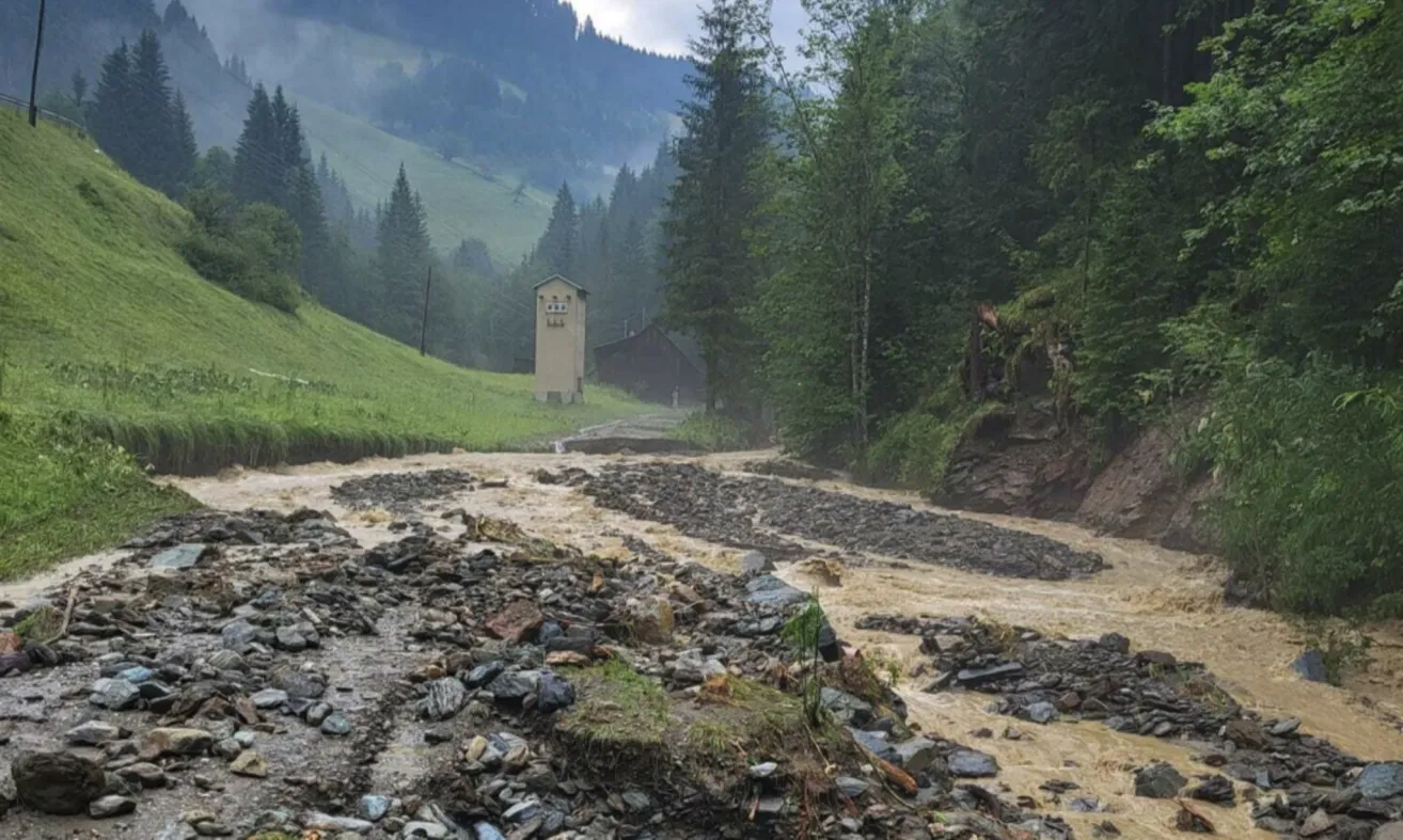 A river with muddy water is flowing through a mountain area. Rocky and grassy areas are on both sides. In the distance, a small house and a mountain are visible.