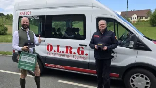 Two men stand next to a white van labeled O.L.R.G. Katastrophenhilfsdienst. One man gives a thumbs-up, while the other holds a hat.