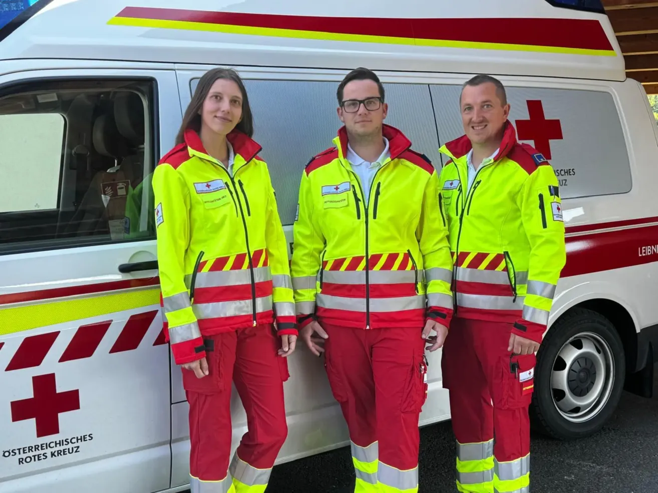 Drei Personen in Warnkleidung, zwei Männer und eine Frau, stehen neben einem Krankenwagen. Sie lächeln und scheinen für ein Foto zu posieren.
