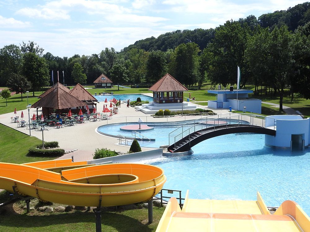 A water park with several pools, a slide, and a bridge. Umbrellas and chairs are set up around the pools.