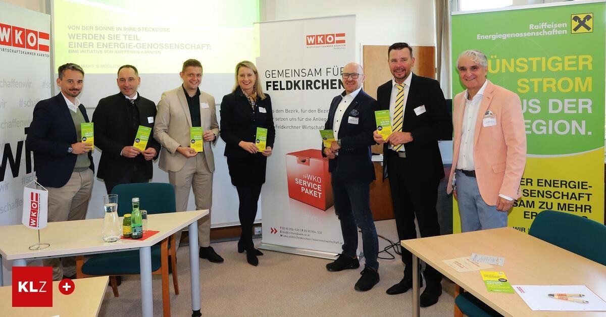 Eine Gruppe von Menschen steht vor einem Banner. Sie halten Broschüren in den Händen und scheinen für ein Foto zu posieren. Hinter ihnen stehen zwei Tische mit einigen Gegenständen darauf. Die Umgebung scheint ein Büro zu sein.