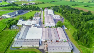 Aerial view of an industrial complex surrounded by green fields and trees, with several buildings and a road leading to it.