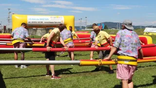 A group of people are playing an obstacle course on a sunny day. They are wearing yellow vests and holding onto a red and yellow tube. A man on the right is wearing a hat and a bracelet.