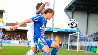 Zwei Fußballspieler sind mitten in einem Spiel. Ein Spieler in Blau versucht, den Ball zu schießen, während der andere in Weiß verteidigt.