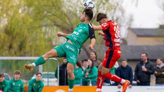 Zwei Fußballspieler, einer in Grün und einer in Rot, springen, um den Ball zu köpfen. Zuschauer beobachten hinter einem Zaun.