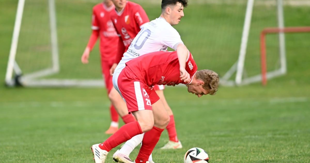 Two soccer players are in a duel for the ball. One player is wearing a red jersey and the other a white jersey. A third player stands in the background.