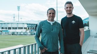 Two men stand on a balcony overlooking a stadium. One wears a teal shirt, the other a black shirt with a logo. Behind them is a field and stadium lights.
