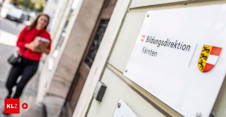 Ein weisses Schild an einer Gebäudewand mit dem Text 'Bildungsdirektion Kärnten' und einem roten Pfeil nach links. Eine verschwommene Figur in Rot geht auf dem Bürgersteig vorbei.
