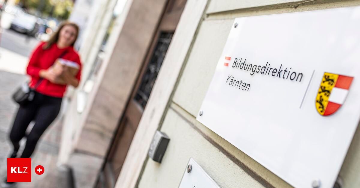 Ein weisses Schild an einer Gebäudewand mit dem Text 'Bildungsdirektion Kärnten' und einem roten Pfeil nach links. Eine verschwommene Figur in Rot geht auf dem Bürgersteig vorbei.