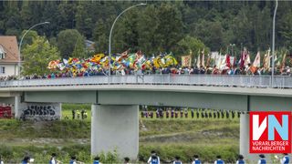A group of people stands on a bridge waving flags, while others are lined up on the grass below. Trees and a building are in the background.