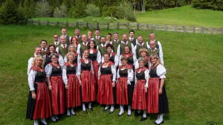 Ein Chor von Menschen in traditioneller deutscher Kleidung, mit roten und schwarzen Kleidern, weißen Blusen und grünen Westen, posiert für ein Gruppenfoto auf einer grasigen Wiese.