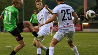 Three soccer players on the field. One player is wearing a white jersey with the number 25. Another player is wearing a green jersey. The player in the middle is kicking the ball.