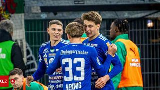 Drei junge Fußballer in blauen Trikots feiern ein Tor, lächeln und umarmen sich. Der Spieler mit der Nummer 33 steht in der Mitte. Hinter ihnen beobachtet ein weiterer Spieler in Grün.