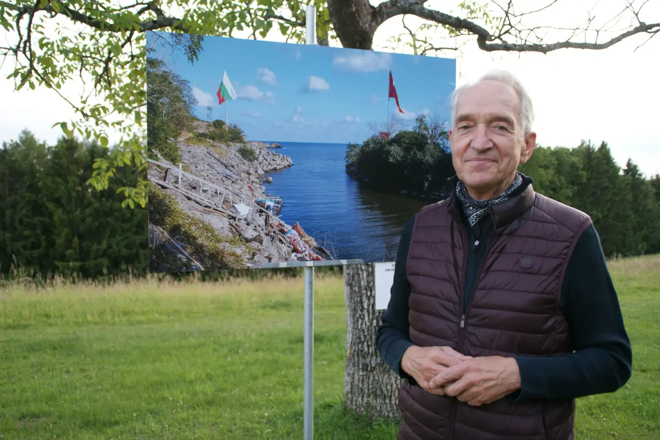 An older man stands in front of a large photograph. It shows a rocky coast with two flags. He wears a brown vest and looks serious.