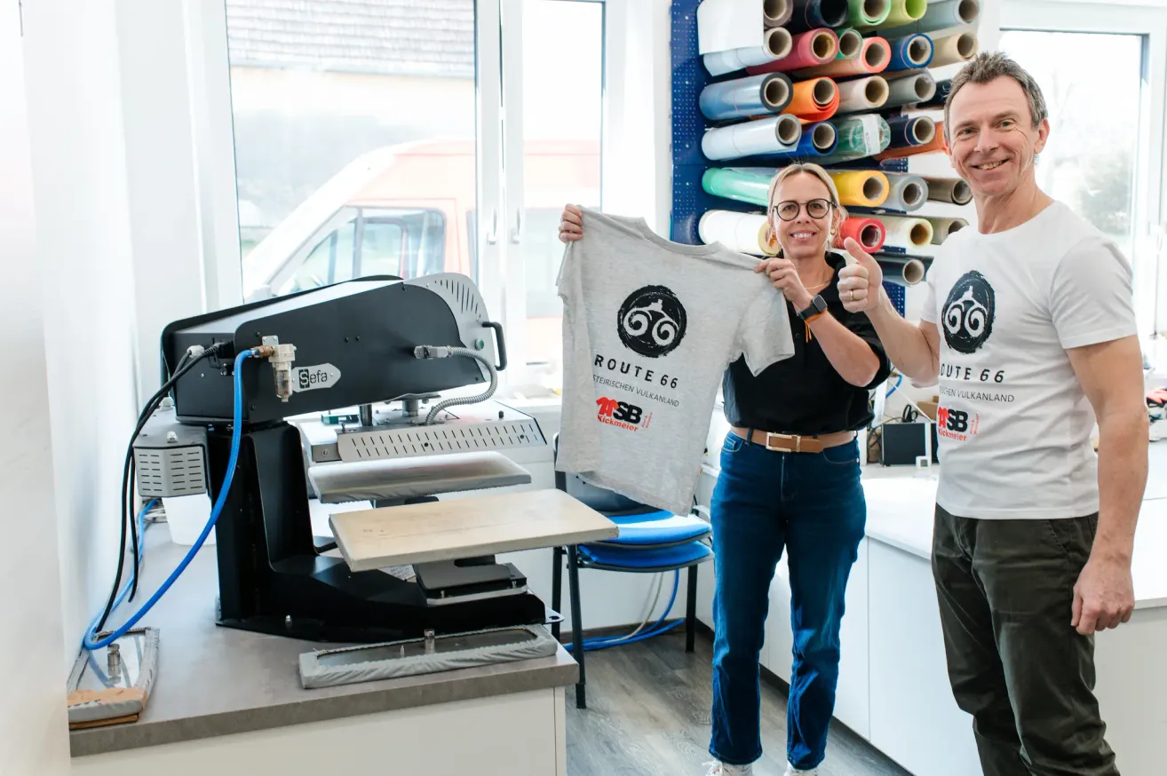 Two people stand in a room with a screen printing machine and a wall of colorful spools. The woman holds a gray shirt with Route 66 written on it.