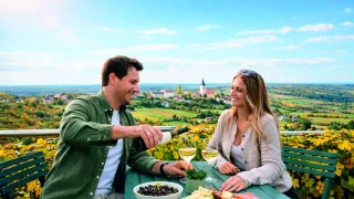 A couple enjoys a meal at an outdoor restaurant with a scenic view. The man pours wine, and they have cheese and olives on the table.
