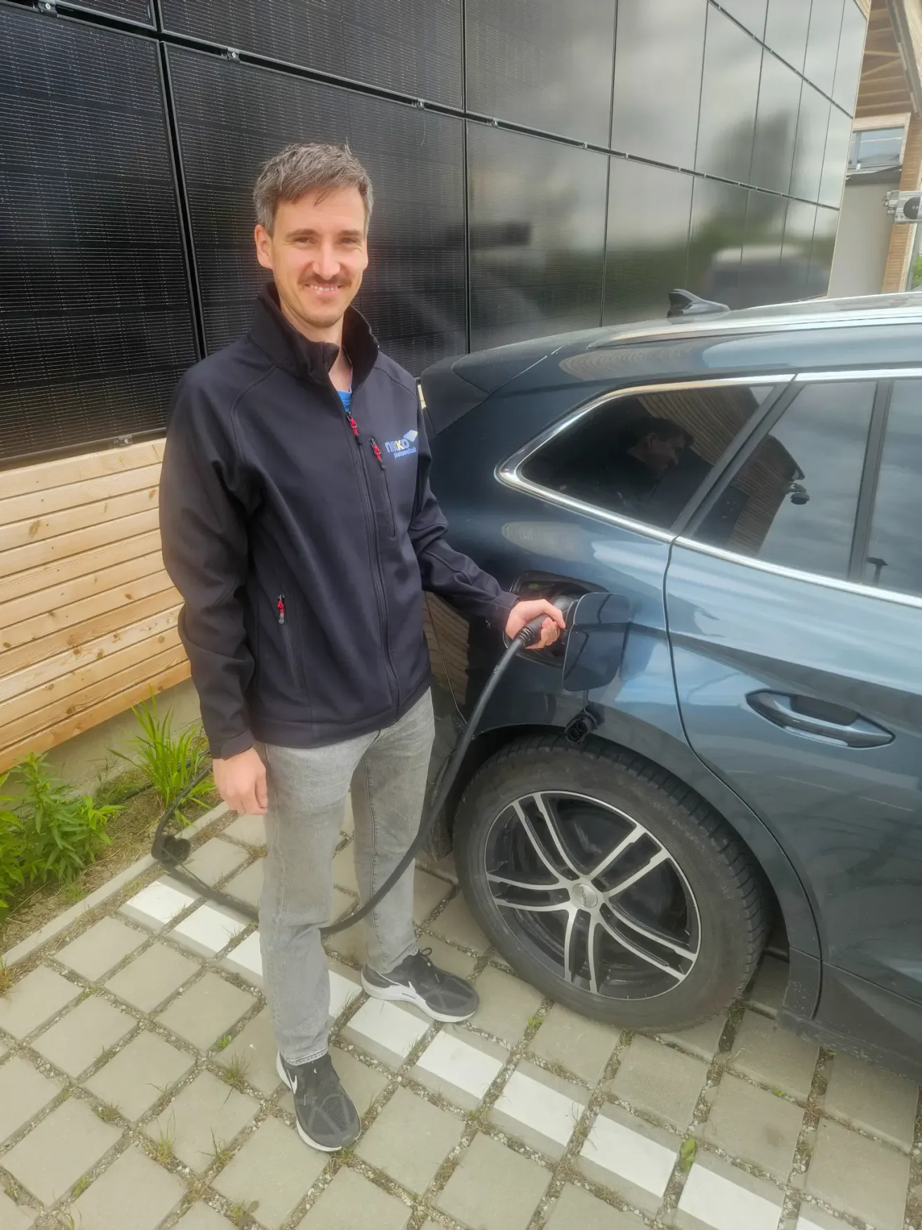 A man is standing beside a gray car and filling it with fuel. He is smiling and seems to be posing for a photo.