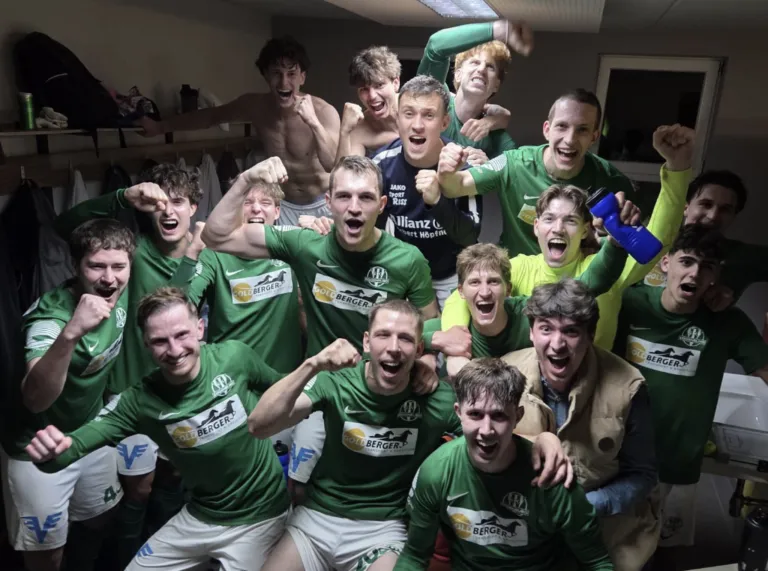 A group of soccer players celebrate in a locker room, wearing green jerseys with sponsor logos. They are cheering and raising their arms, some holding a blue bottle. The room has a white ceiling and a window on the right.