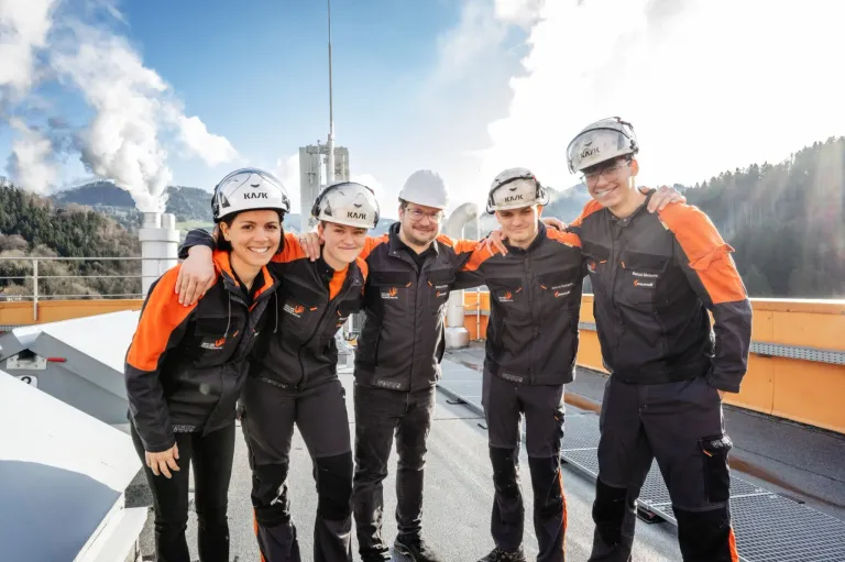 Fünf Personen in Schutzkleidung posieren vor einer industriellen Umgebung für ein Foto, mit zwei Männern an jeder Seite einer Frau.