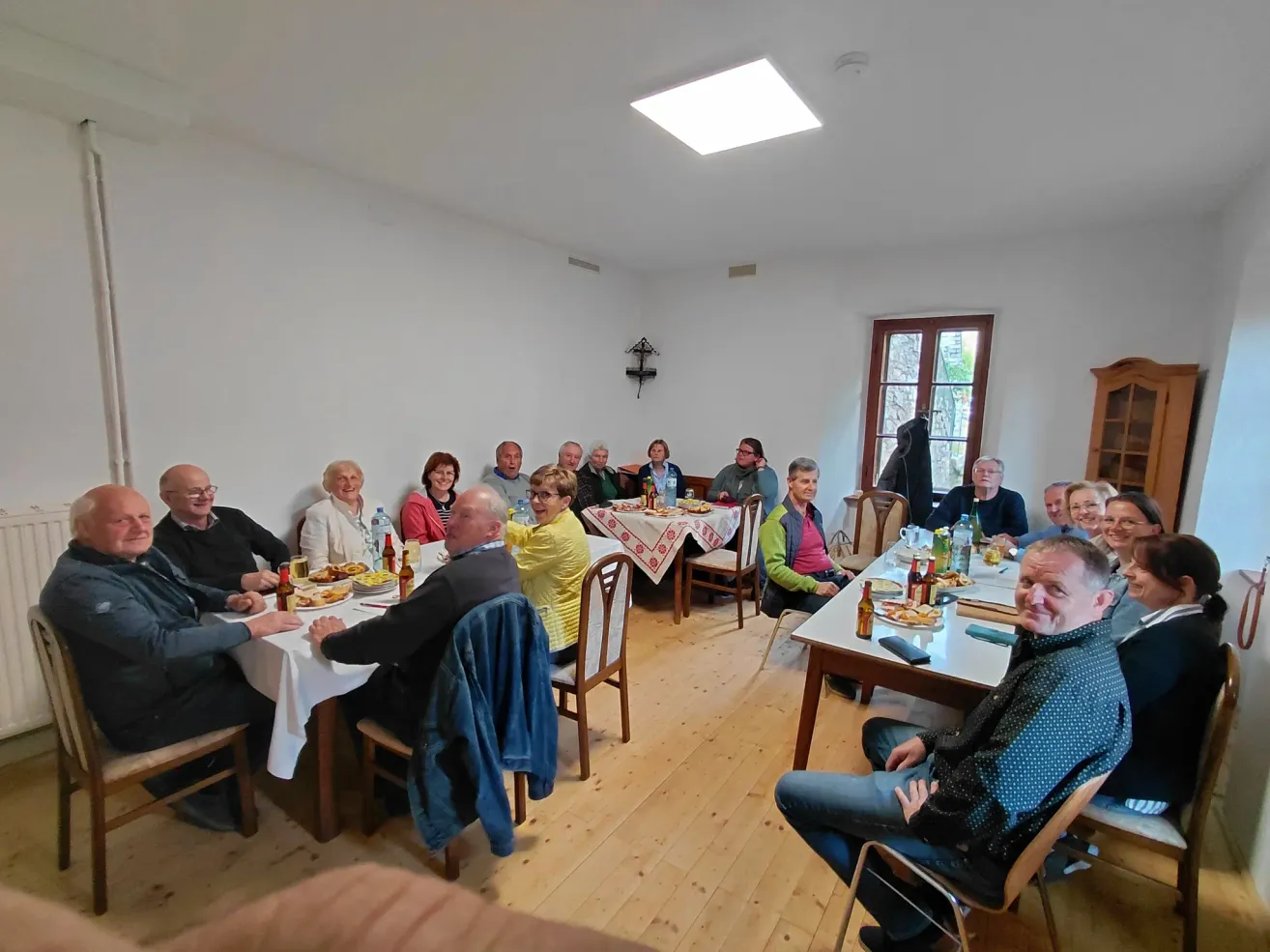 Eine Gruppe von Menschen sitzt an einem Tisch in einem Raum und genießt eine Mahlzeit. Einige tragen Brillen, und Flaschen und Teller sind auf dem Tisch.