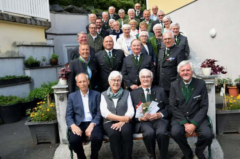 Eine Gruppenaufnahme von älteren Menschen in Uniformen, die Urkunden und Blumen halten, sitzen auf einer Bank vor einem Gebäude.