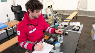 A man in a red jacket is writing on paper while holding a pen and surrounded by scientific equipment.