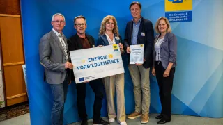 Five people stand in front of a blue wall with the word enw. Four of them hold certificates and one holds a board that says Energie Vorbildgemeinde.