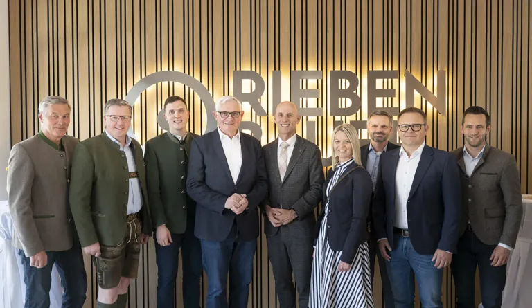 Sieben Menschen stehen vor einer Wand mit dem Wort 'Riebes'. Sie sind alle formell gekleidet, tragen Brillen und lächeln für die Kamera. Der Hintergrund ist holzfarben mit vertikalen Streifen.