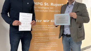 Two men stand next to each other holding certificates, in front of a banner with a logo and text about St. Rupert. They are both wearing jackets and jeans.