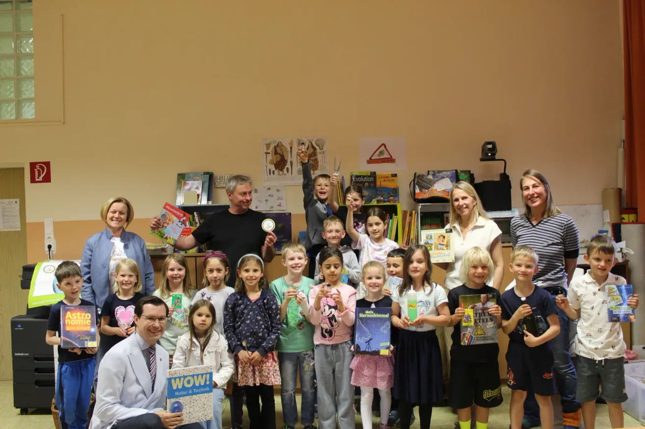 Eine Gruppe von Kindern und Erwachsenen posiert mit Büchern in einem Klassenzimmer für ein Foto. Ein Mann hebt die Hand, und einige Kinder halten Bücher.