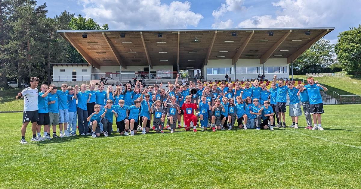 Eine große Gruppe von Kindern in blauen Uniformen posiert für ein Foto auf einem Rasenfeld, mit einer überdachten Tribüne im Hintergrund.