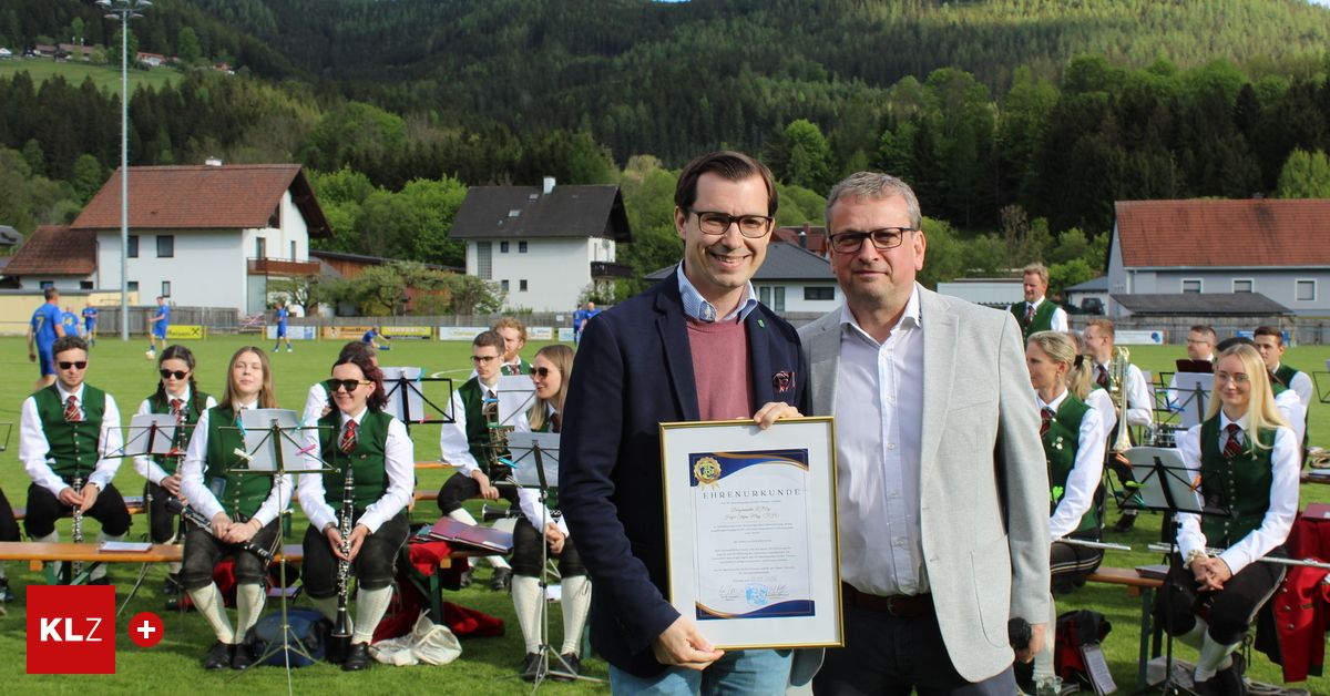 Zwei Männer stehen lächelnd auf einem Feld und halten ein Zertifikat. Hinter ihnen sitzen Musiker auf dem Gras und spielen Instrumente. Häuser sind im Hintergrund, mit einem Bergrücken in der Ferne.