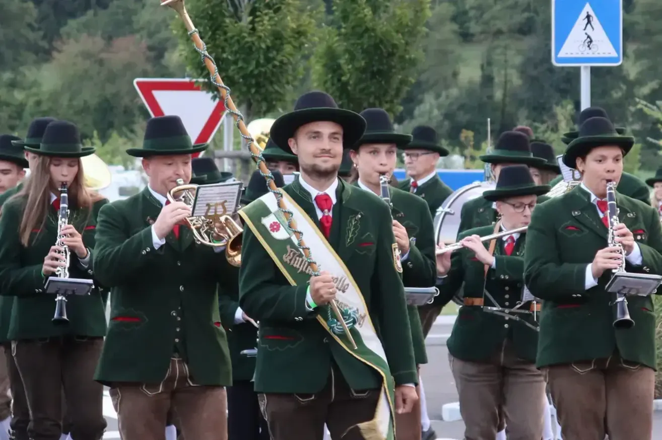 A marching band dressed in green uniforms plays instruments, including trumpets and flutes, in an outdoor parade.