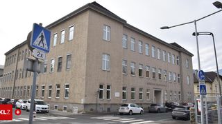 Ein großes mehrstöckiges Gebäude mit zahlreichen Fenstern. Ein Fußgängerüberweg-Schild ist sichtbar. Mehrere Autos sind vor dem Gebäude geparkt.