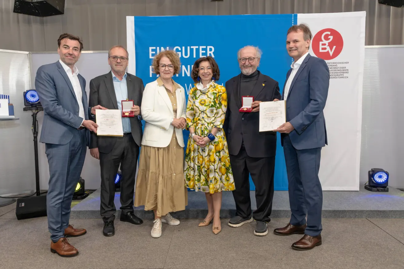 Six individuals pose with awards at a formal event. They are in front of a banner with the text 'Ein Guter'. Each person holds a certificate. The man on the far right smiles.
