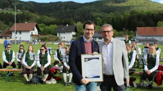 Zwei Männer stehen vor einem grünen Hintergrund und halten ein Zertifikat in den Händen. Hinter ihnen sitzt eine Band auf einer Bank. Im Hintergrund ist eine Berglandschaft zu sehen.