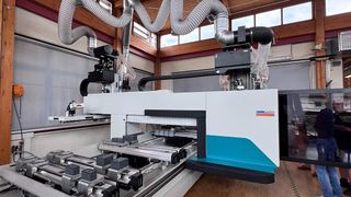 A large industrial machine is in a factory with large windows. It has many pipes and tubes attached. The machine is white and blue with black accents and the word 'HANDL' is on the side.