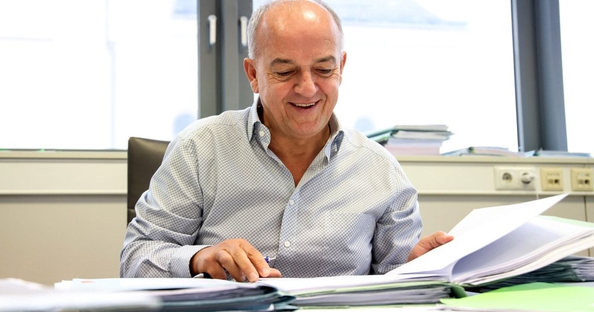 A man in a checkered shirt is sitting at a desk, smiling and holding a pen. Papers and books surround him, suggesting he is engaged in work.