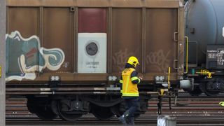 A worker in a yellow safety vest inspects a brown train car with graffiti on the side. The train car has a white panel with text and a circular sign indicating no smoking.