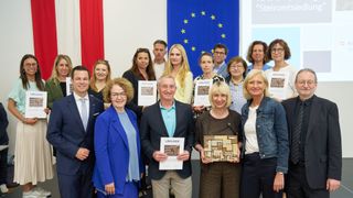 A group of people are holding certificates in front of a flag and banner. The man in the middle is smiling and holding a certificate. They are all wearing formal attire and glasses. The flag has stars and is blue. The banner has a text and images.