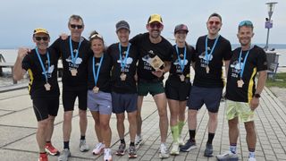 A group of six people, all wearing black shirts and shorts, stand together for a photo. They have medals around their necks and are smiling. The ground beneath them is brick.