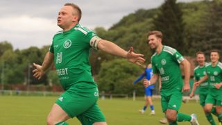 Two soccer players in green uniforms run on a field. One points forward, while another smiles behind. Trees and a fence are in the background.