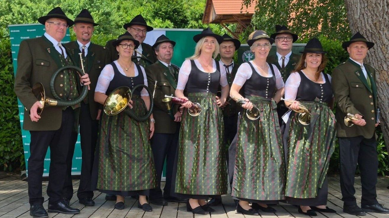 A group of people in traditional Bavarian clothing are standing together. They are holding brass instruments and smiling for a photograph.
