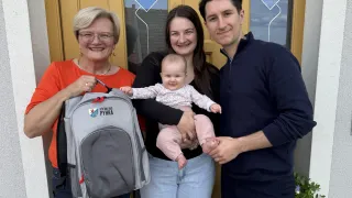 A woman, a man, and a baby, all smiling and posing for a photo. The woman holds a backpack with a logo and the text 'Ich bin aus PYHRA'. Behind them, there's a door with a glass window.